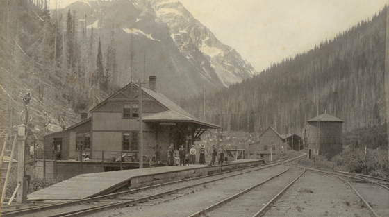 Photo en noir et blanc de six passagers qui attendent sur le quai en bois d'une petite gare de train en été. Chaîne de montagnes en arrière-plan.