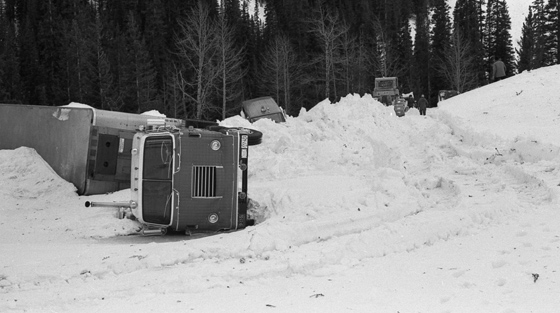 Black and white photograph of truck thrown on to its side after being tossed through the air by avalanche. Rescuers in background.