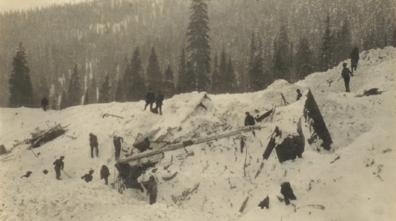 Black and white photograph of 10+ rescuers on site of avalanche, which shows a rotary snow plough pushed off the railway tracks and uphill.