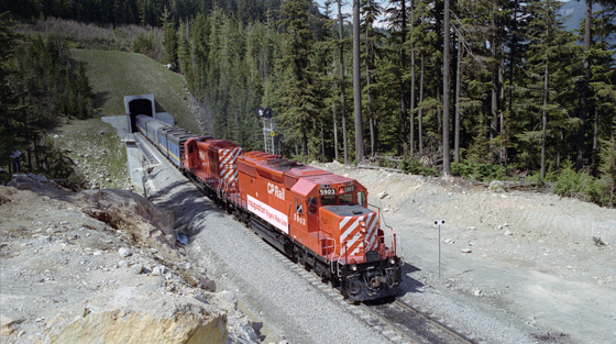 Colour photograph of train with red engine as it emerges from mountain tunnel in summer.