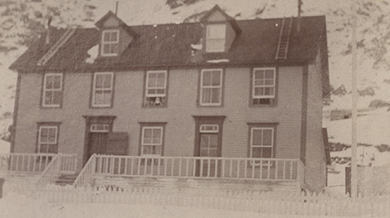 Photo en noir et blanc d'une maison en bois à deux étages en hiver et derrière elle, une pente montagneuse escarpée.