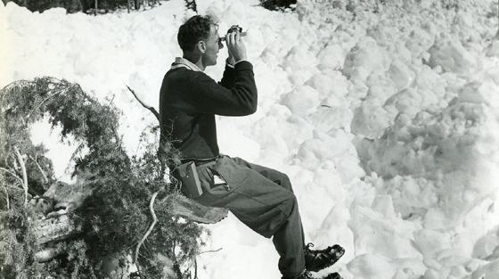 Black and white photograph of man sitting on log in snow looking up a slope with specialized equipment. 