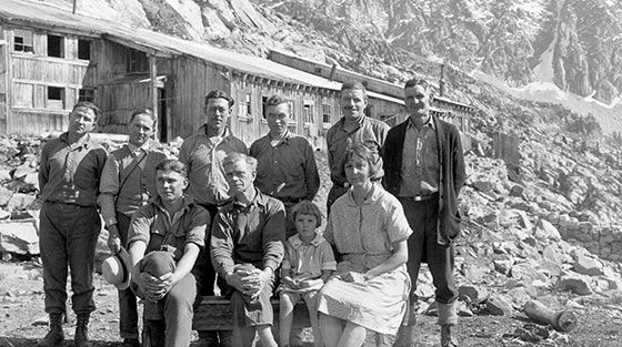 8 men, 1 woman and 1 child pose for black and white photograph with building in background, in summer.
